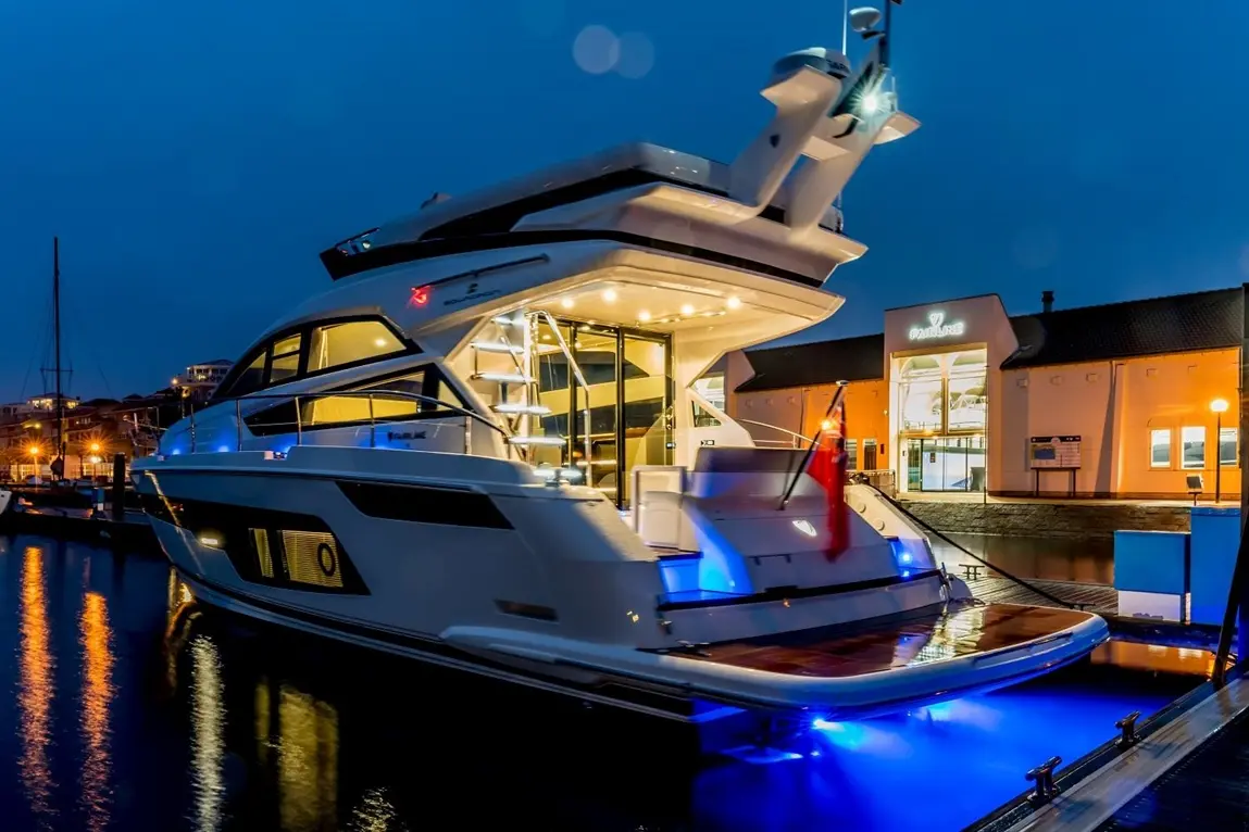 Luxury yacht docked at night, illuminated by blue lights, with a modern marina building in the background.